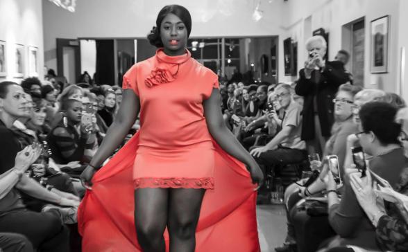 black and white photo in the galley of SPNN. The shot is of a runway model walking down the hallway with people sitting on both sides. Her dress is the only thing in color and it is red. 