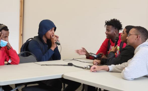 five students around a microphone. One with headphones on creating a podcast