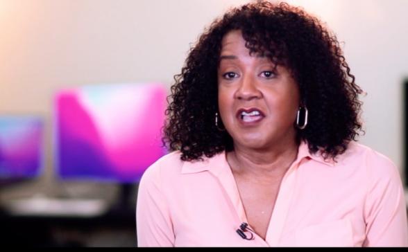 This is a still image from the video. It features Allison in a pink shirt sitting in front of computers. The computers are soft focused. 