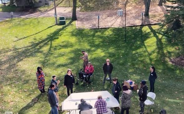 an outdoor shot of several people gathered in a park. Two are sitting around a table and the others are standing in small groups talking. 