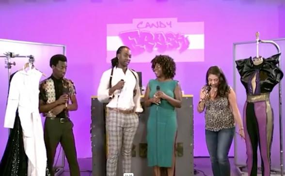 four people are standing in front of a pink wall that says Candy Fresh. There are outfits hanging on either side of the four people (on display). 