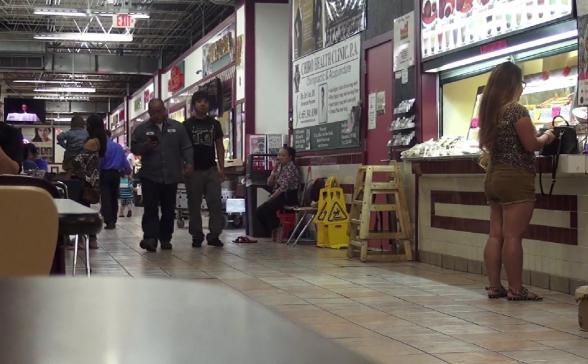A shot of a busy food court with people walking through it and standing by the restaraunts ordering their food. 