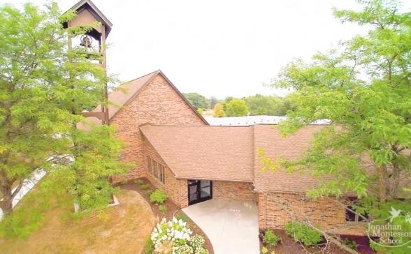 An ariel shot of the Jonathan Montessori School.  There is a large brick building  with a bell tower . There is lots of trees and landscaping around the building. 