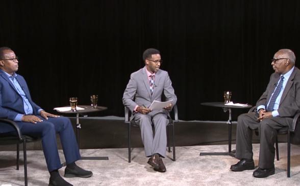 Three Somali men are sitting in front of a black curtain. This is a still from the show. 