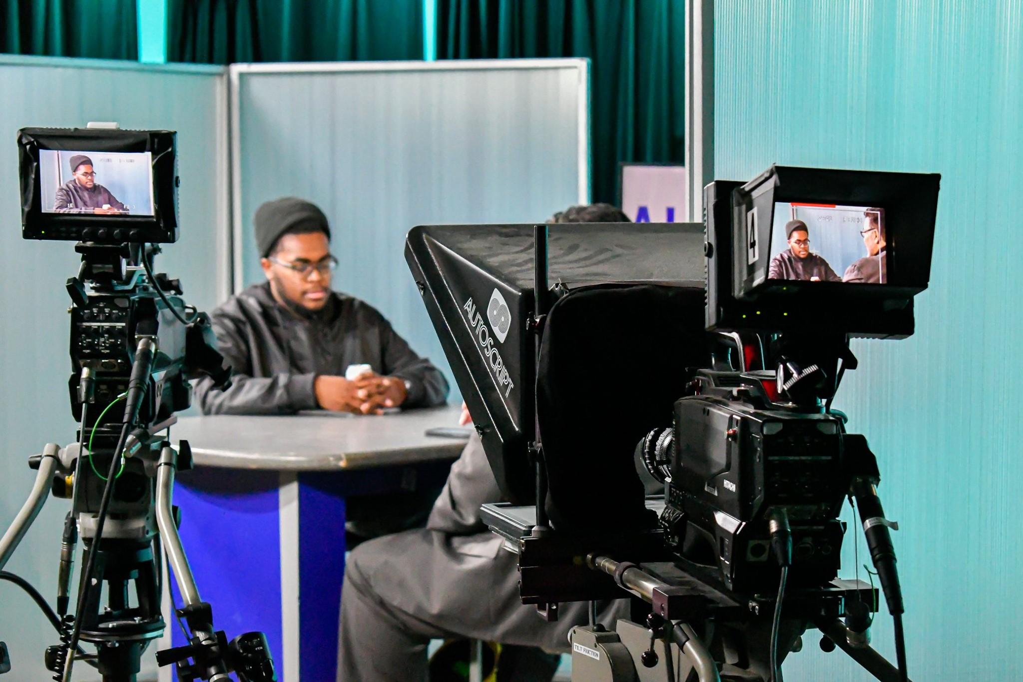 shot of the studio with a teal background with two frosted panels and a guy sitting at a desk. In the foreground there are two studio cameras and you can see their viewfinders. In the viewfinders you can see the one shot of the guest and in the other you can see a two shot of the guest and host.