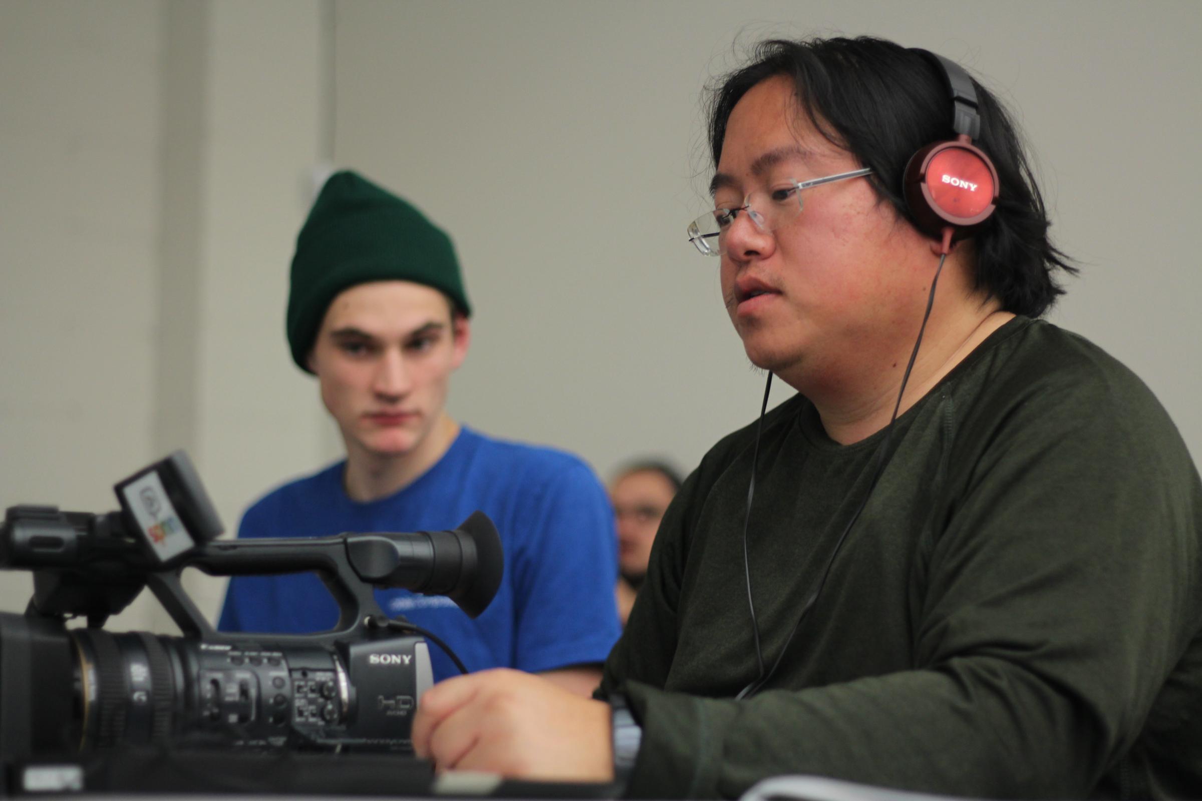 a photo of SPNN's camera class. In the forefront is a guy in green with headphones and an AX2000 in the background is a guy in a blue shirt with a stocking hat on. 