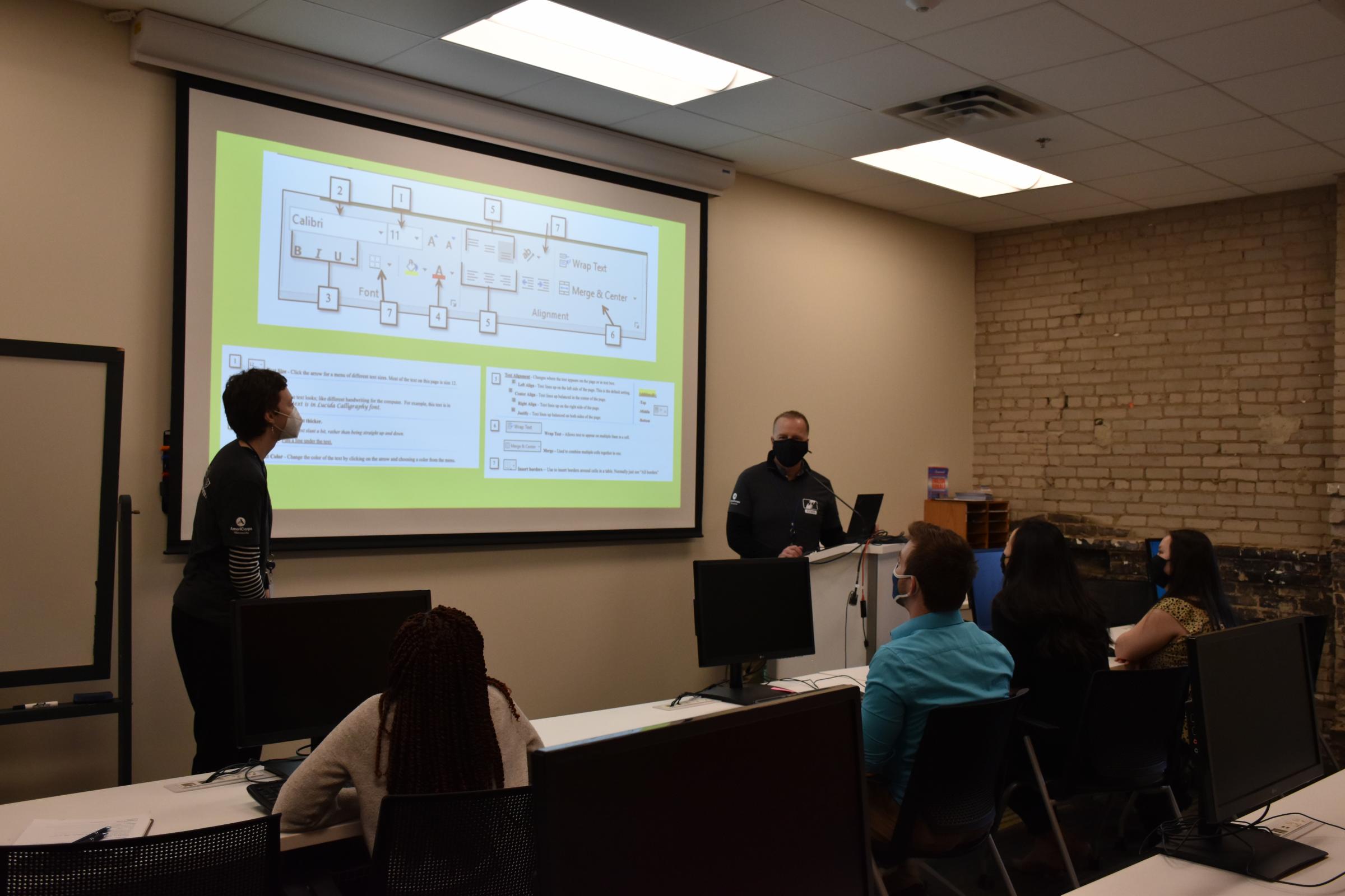 Two CTEP members instruct using a projector and Powerpoint at the front of a classroom with students.